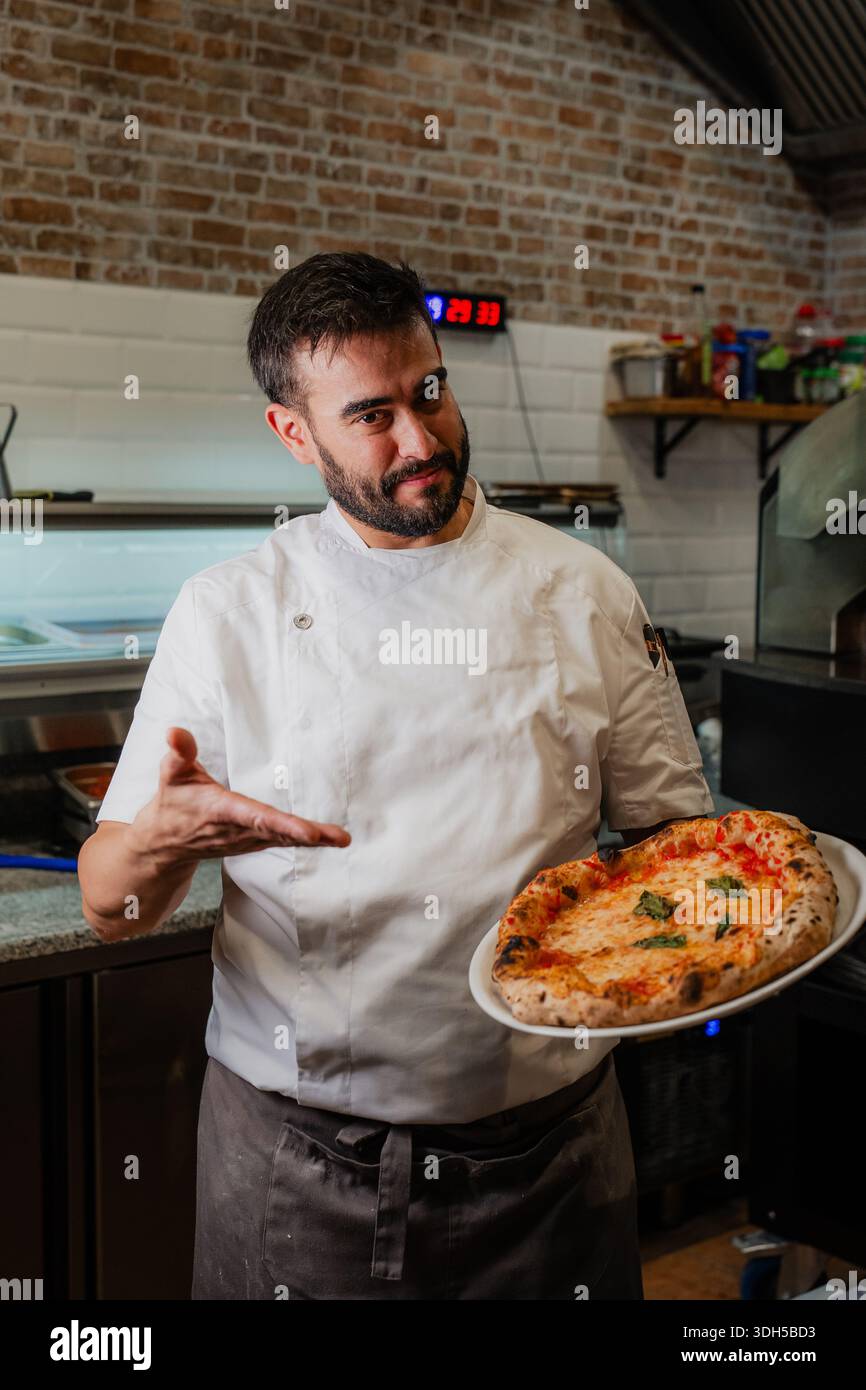 Un pizzeria qualifié présente fièrement une délicieuse pizza napolitaine, fraîche du four. Il porte une tenue de chef blanche, symbolizin Banque D'Images