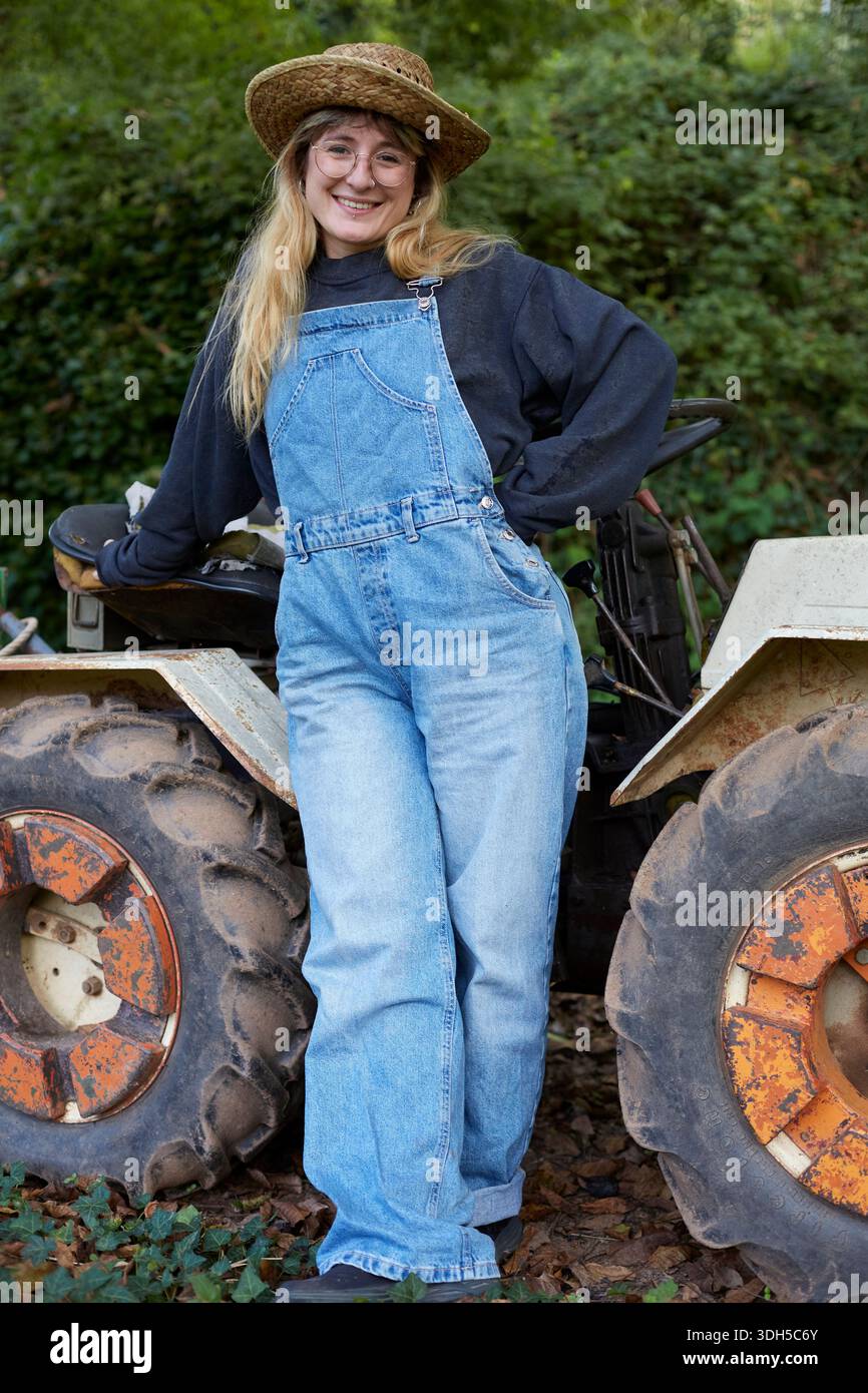 Une femme en salopette en denim et un chapeau de paille se tient à côté d'un tracteur, posant avec confiance dans le jardin verdoyant et luxuriant. Elle sourit chaleureusement, capturant l'essen Banque D'Images