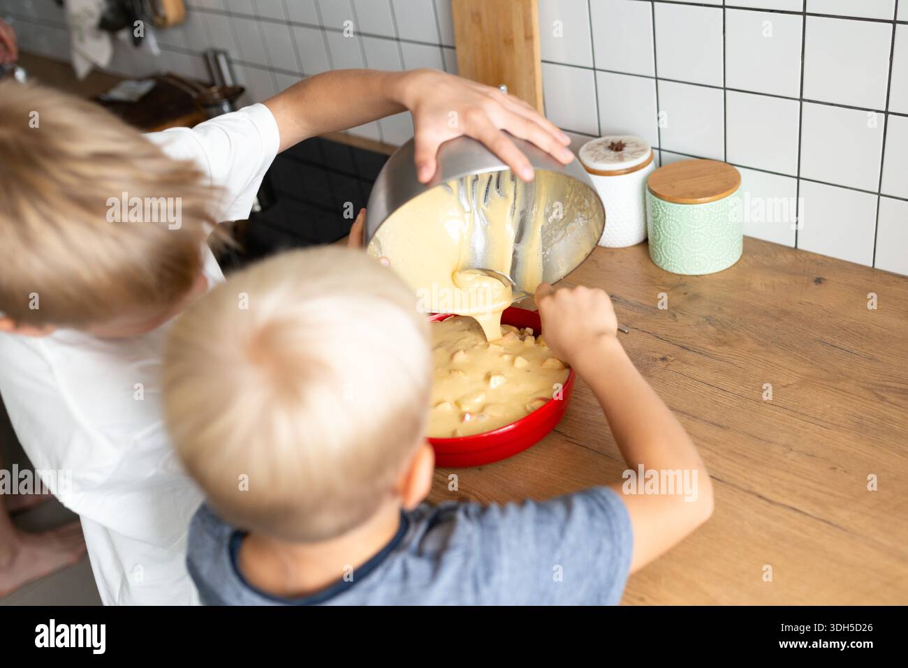 Deux enfants préparent avec impatience une pâte à gâteau dans une cuisine confortable, versant le mélange dans un bol rouge vif. Leur travail d'équipe et leur plaisir soulignent le Banque D'Images
