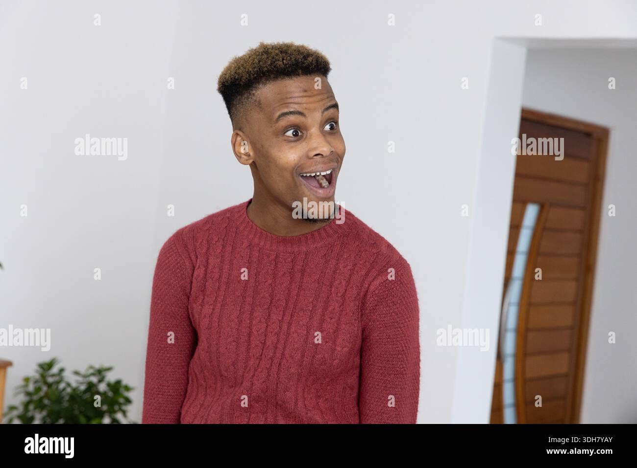 Homme afro-américain debout dans le hall portant un pull rouge, regardant à droite la porte en bois et la plante Banque D'Images