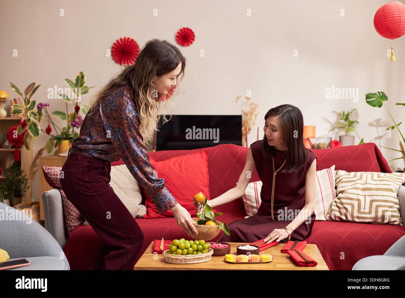 Jeune femme asiatique adulte arrangeant des fruits sur la table, tandis qu'une autre jeune femme asiatique adulte assise sur le canapé atteignant la nourriture pendant la célébration des vacances Têt dans le salon décoré avec des ornements rouges Banque D'Images