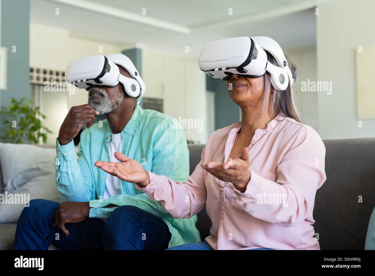Couple senior portant des casques VR blancs assis sur un canapé dans le salon interagissant virtuellement Banque D'Images