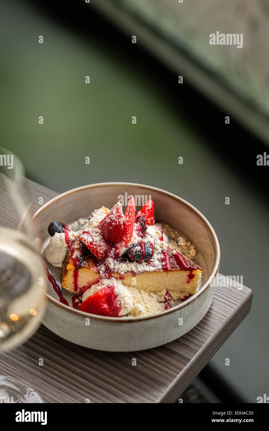 Une délicieuse tranche de cheesecake basque brûlé crémeux servi dans un bol en céramique, garnie de fraises fraîches, de myrtilles et de sucre en poudre. Banque D'Images
