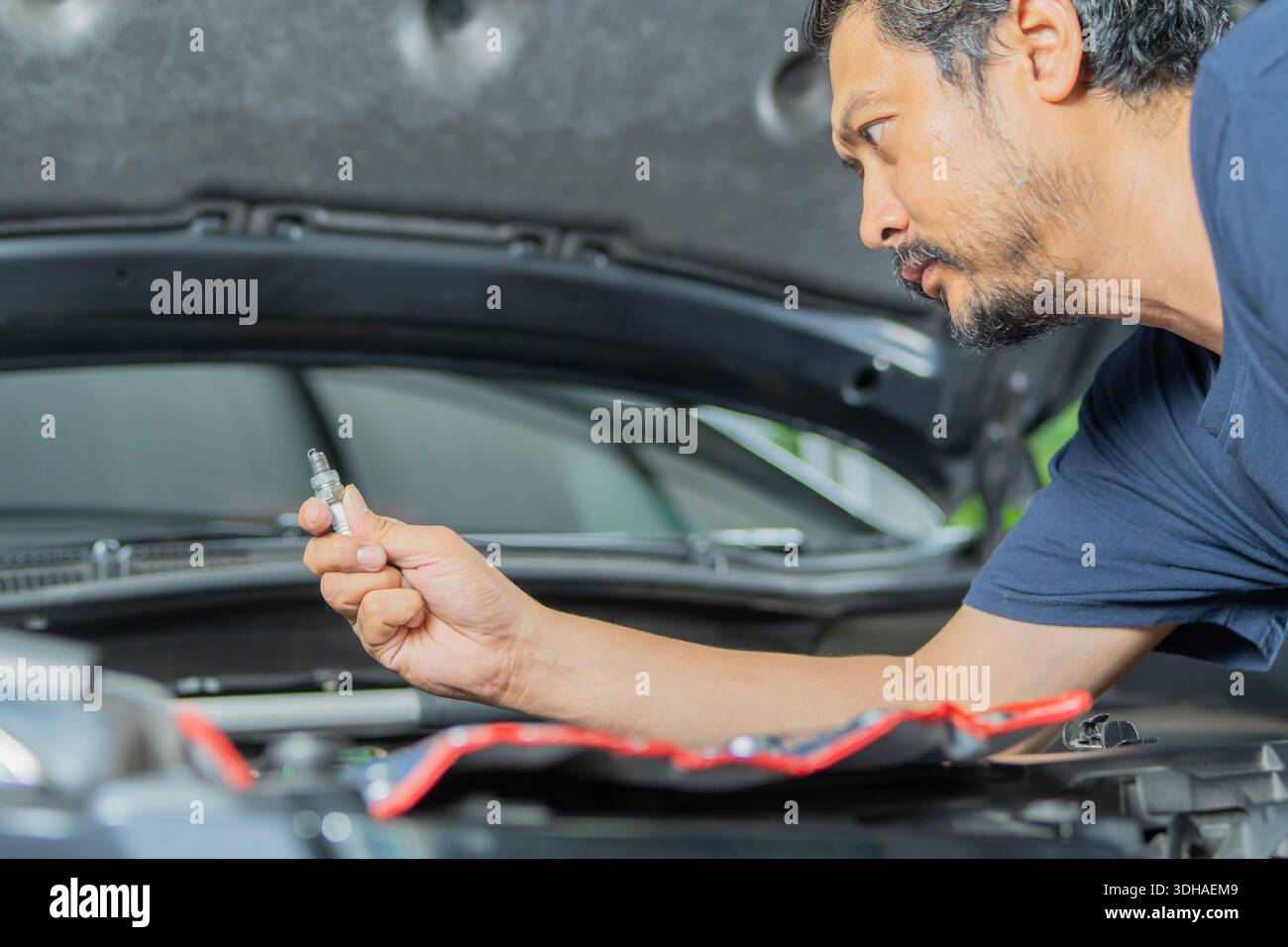 Vue latérale d'un bel homme asiatique avec barbe change de bougies d'allumage dans sa voiture. Remplacement des bougies d'allumage dans la voiture. Banque D'Images