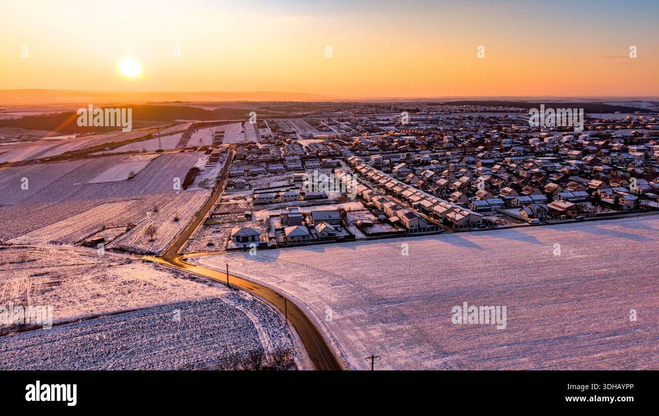 Vue aérienne des terres agricoles et des quartiers résidentiels poussiéreux de neige à Miroslava, comté de Iasi, Roumanie, au lever du soleil, avec des champs vallonnés, routes rurales et Banque D'Images