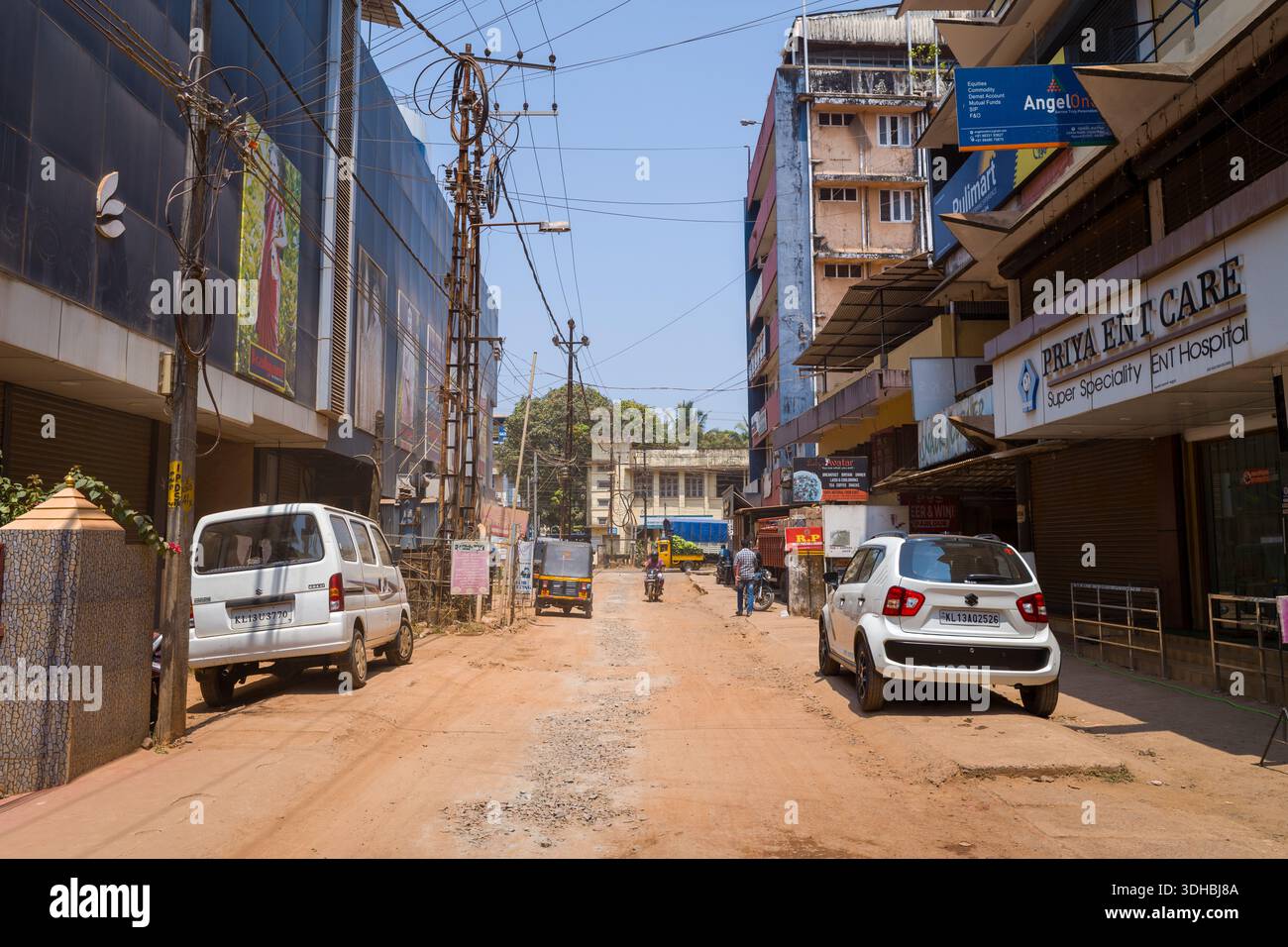 Kannur, Inde - 13 mars 2023 : une rue de terre à Kannur montre des voitures garées, un pousse-pousse automobile, des piétons, des magasins de détail avec des panneaux en anglais et des lignes électriques visibles sous la lumière du jour. Banque D'Images
