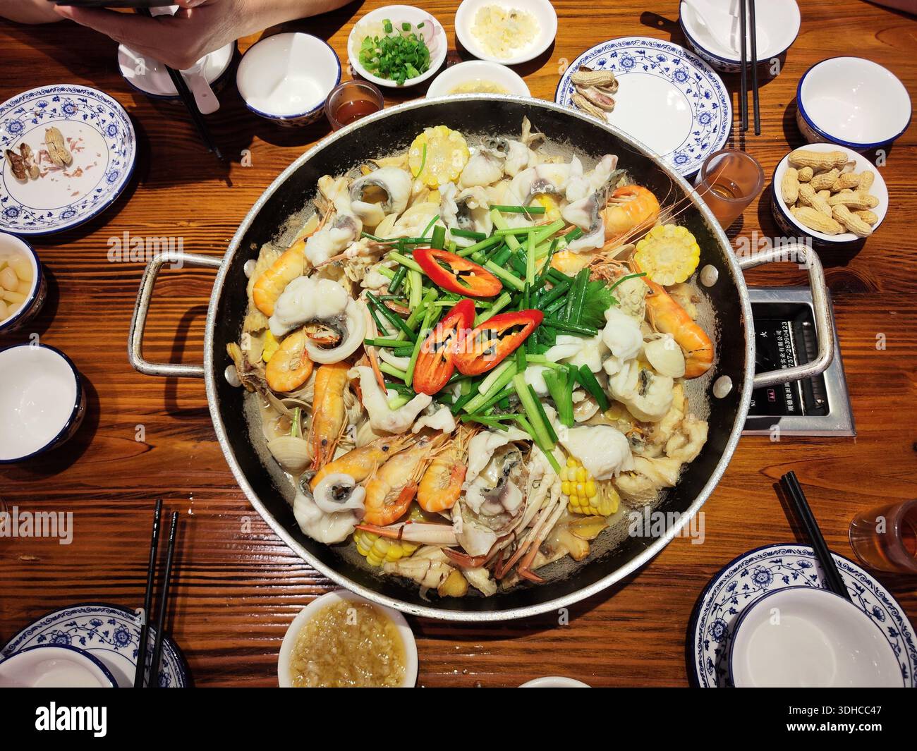 Un repas chaud de fruits de mer vibrant servi dans un grand pot commun, entouré de plats d'accompagnement et de baguettes sur une table en bois. Banque D'Images