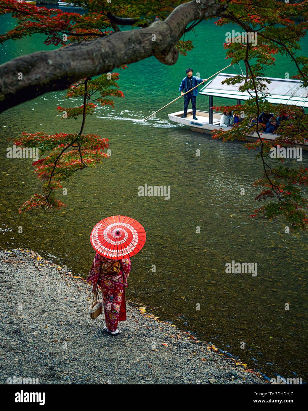 Kyoto, Japon - 18 janvier 2026 : vue d'une femme dans un kimono vibrant et un parasol rouge descendant vers la rivière, tandis qu'un batelier navigue dans le turquo Banque D'Images