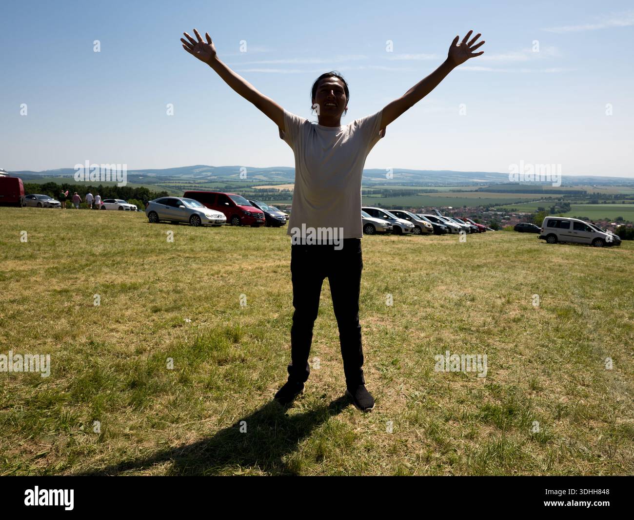 Un homme des années 30 debout les bras levés sur une colline herbeuse, avec une rangée de voitures garées et un large paysage en République tchèque. Banque D'Images