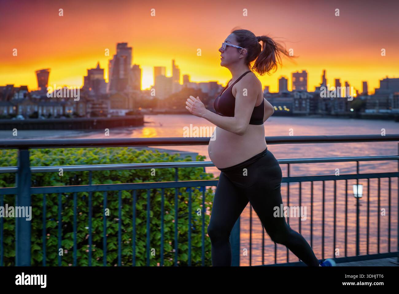 Une femme enceinte en vêtements de sport courant pour l'exercice devant une ligne d'horizon urbaine pendant un coucher de soleil ardent Banque D'Images