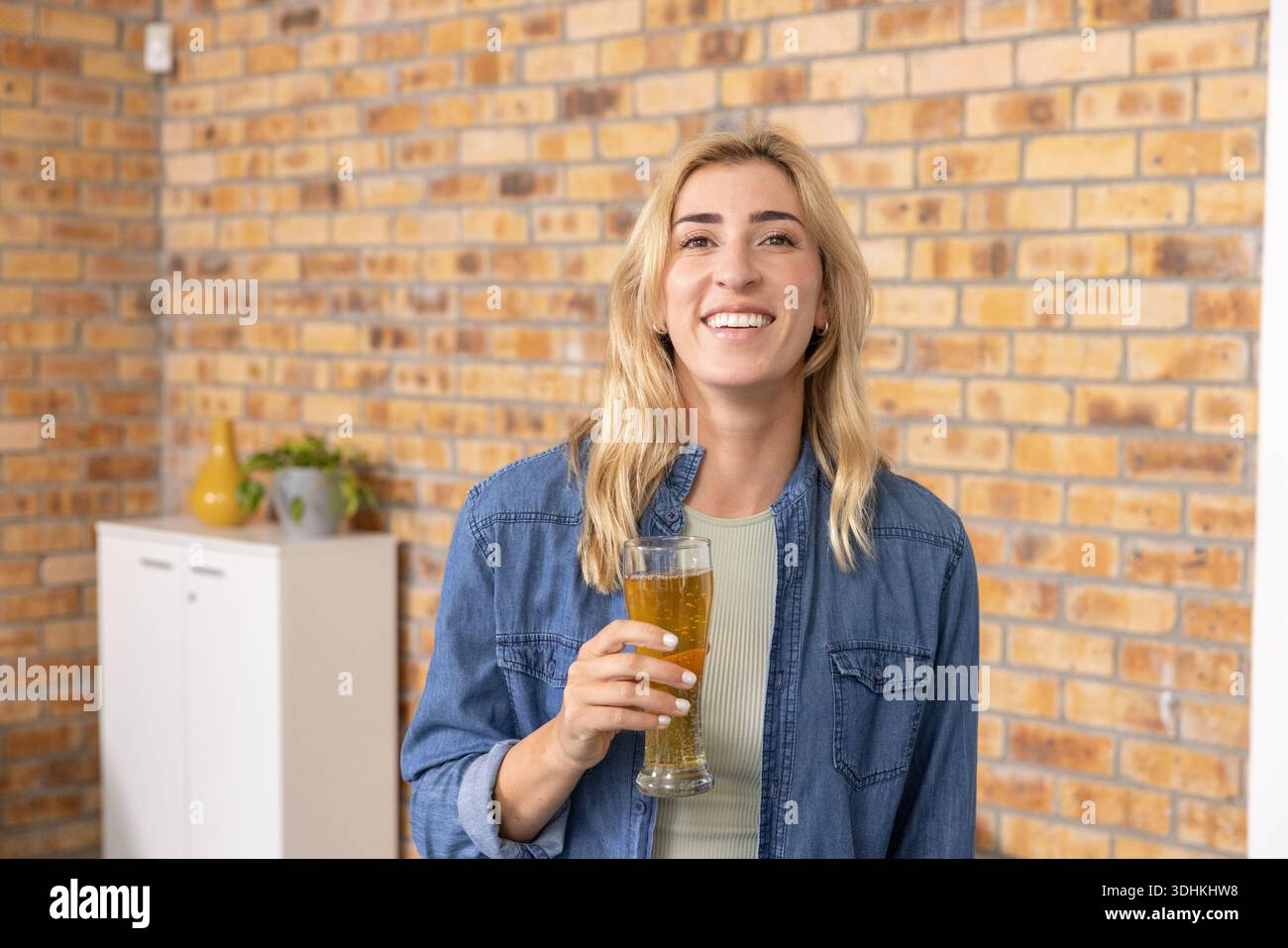 Femme mi-adulte portant une veste en denim tenant le verre de bière contre le mur de briques par armoire, vase Banque D'Images