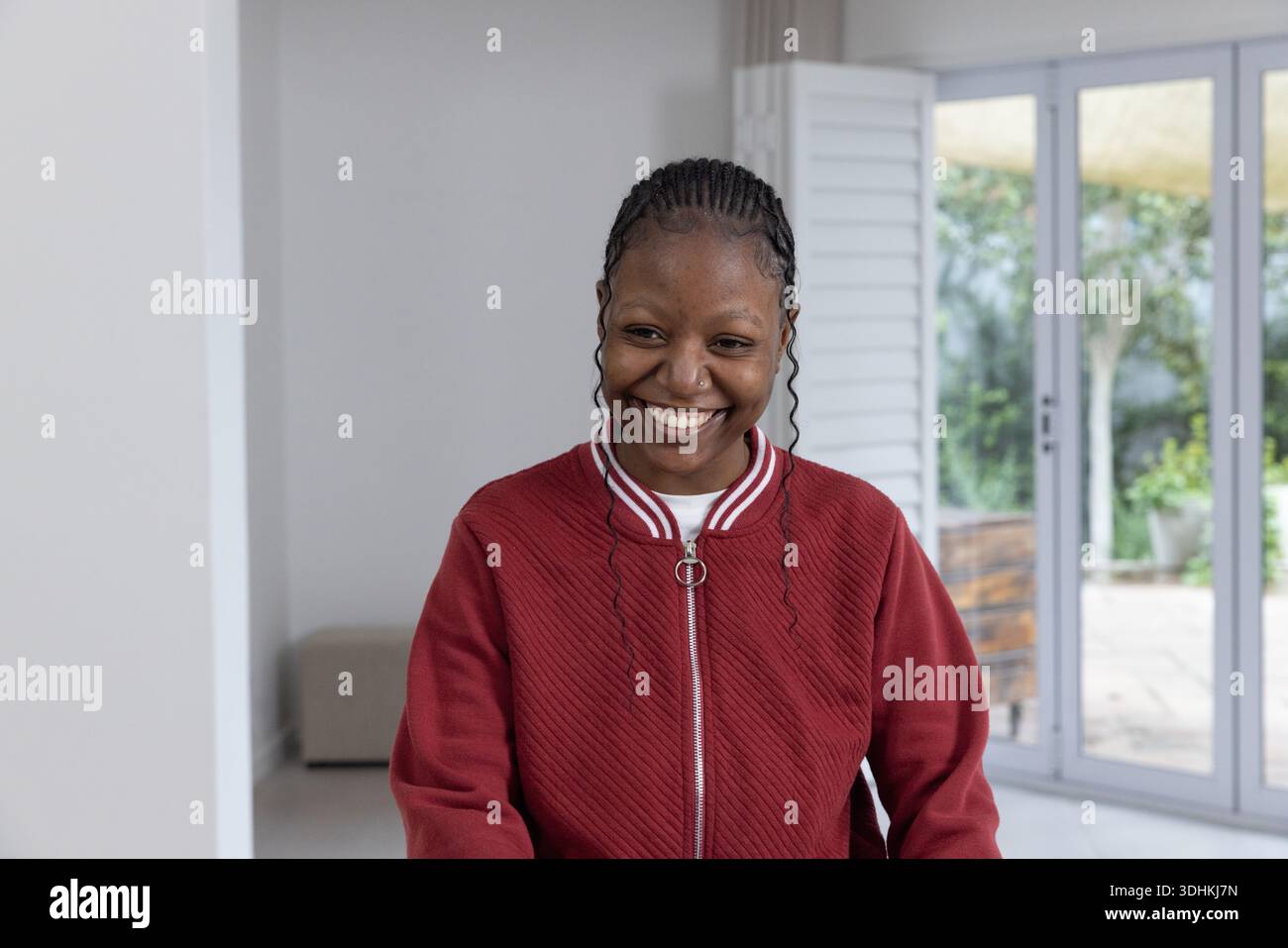 Femme afro-américaine debout et souriante à la maison par des portes coulissantes portant une veste rouge avec des tresses Banque D'Images