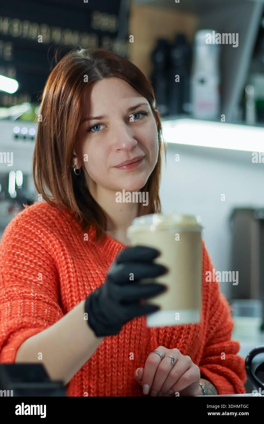 Le barista en tenue orange et gants noirs offre une tasse à café sur le comptoir dans un cadre confortable Banque D'Images