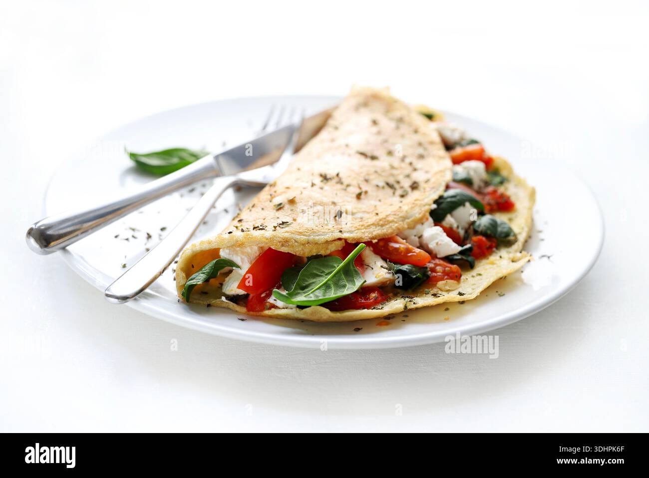 Omelette salée farcie d'épinards frais, de tomates cerises et de fromage sur une assiette blanche, accompagnée d'argenterie. Fond blanc. Plat français. Banque D'Images