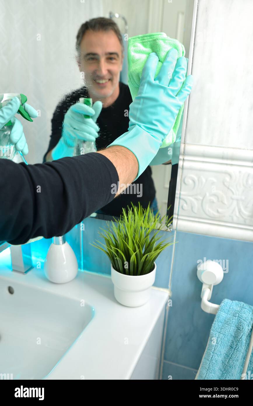 Homme souriant avec des gants en caoutchouc nettoyant le miroir de salle de bain avec un chiffon et vaporisant avec du nettoyant pour vitres. Banque D'Images