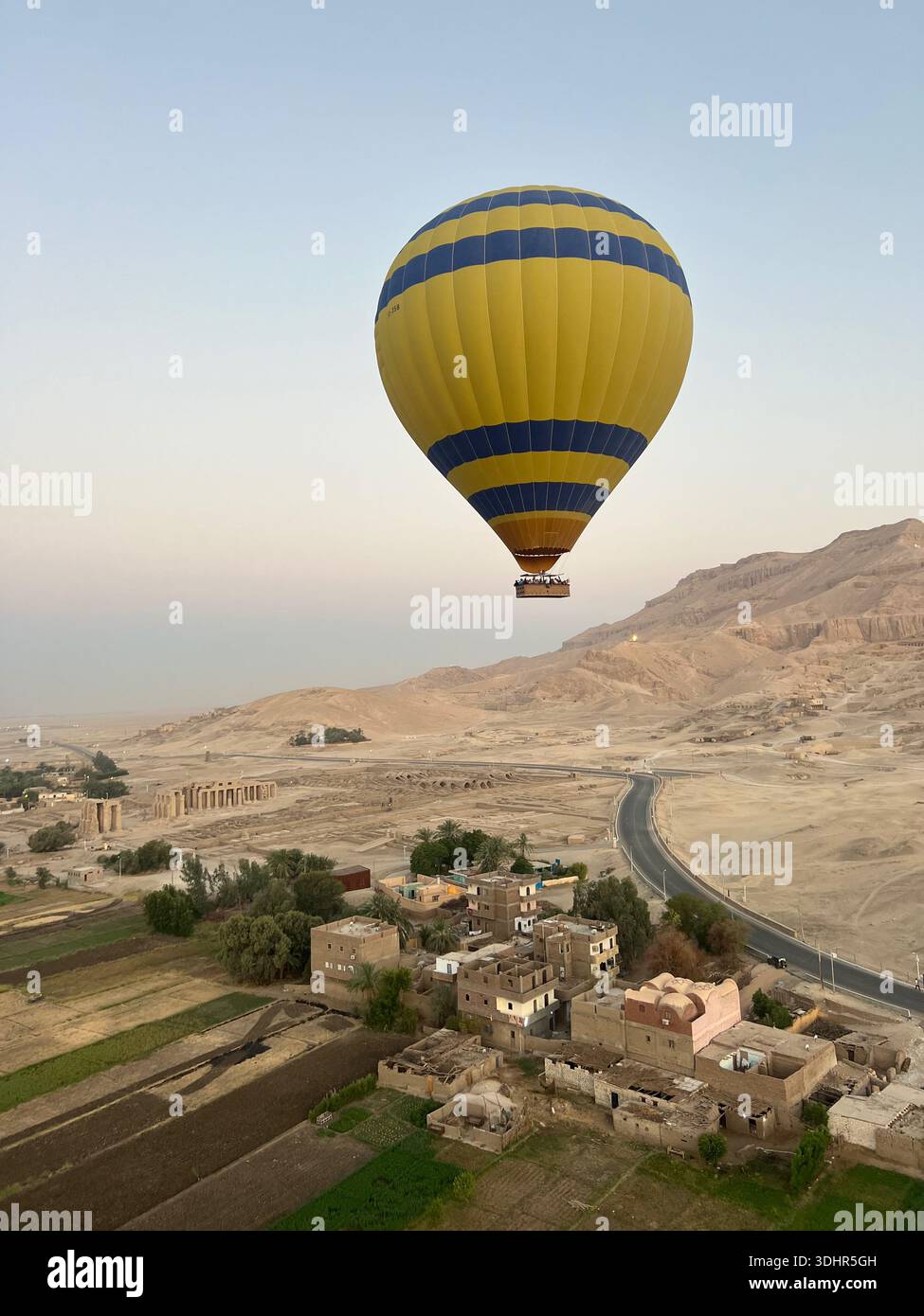 Une montgolfière jaune avec des bandes bleues vole au-dessus des montagnes et des bâtiments résidentiels dans la vallée de Louxor en Égypte - Image de stock capturée avec un smartphone