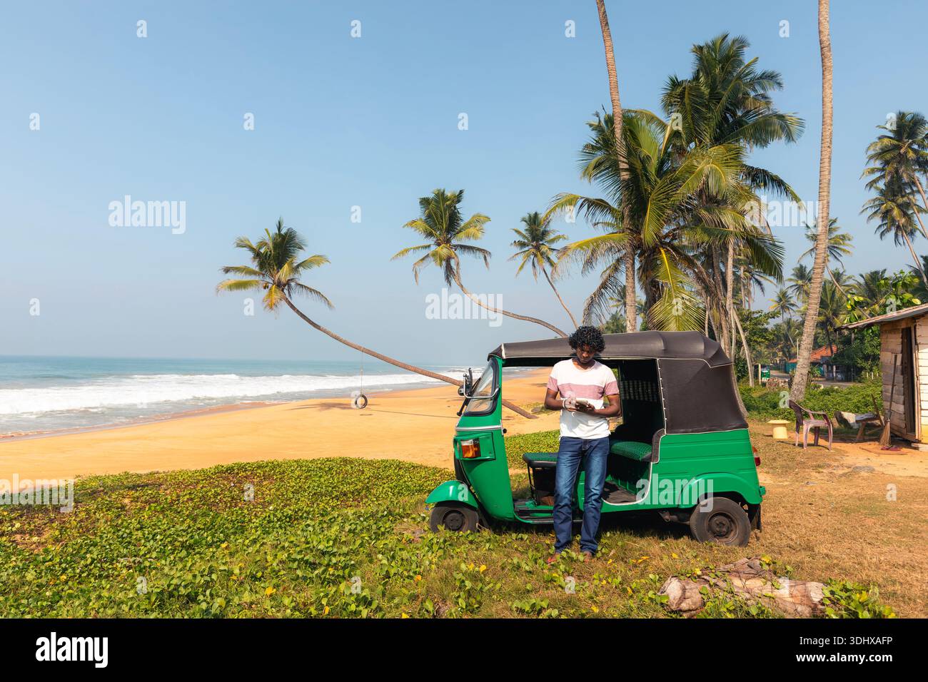 Chauffeur de tuk tuk local attendant à côté du véhicule près de la plage de sable avec des palmiers pendant la journée ensoleillée au Sri Lanka. Transport insulaire authentique, vie quotidienne, Banque D'Images