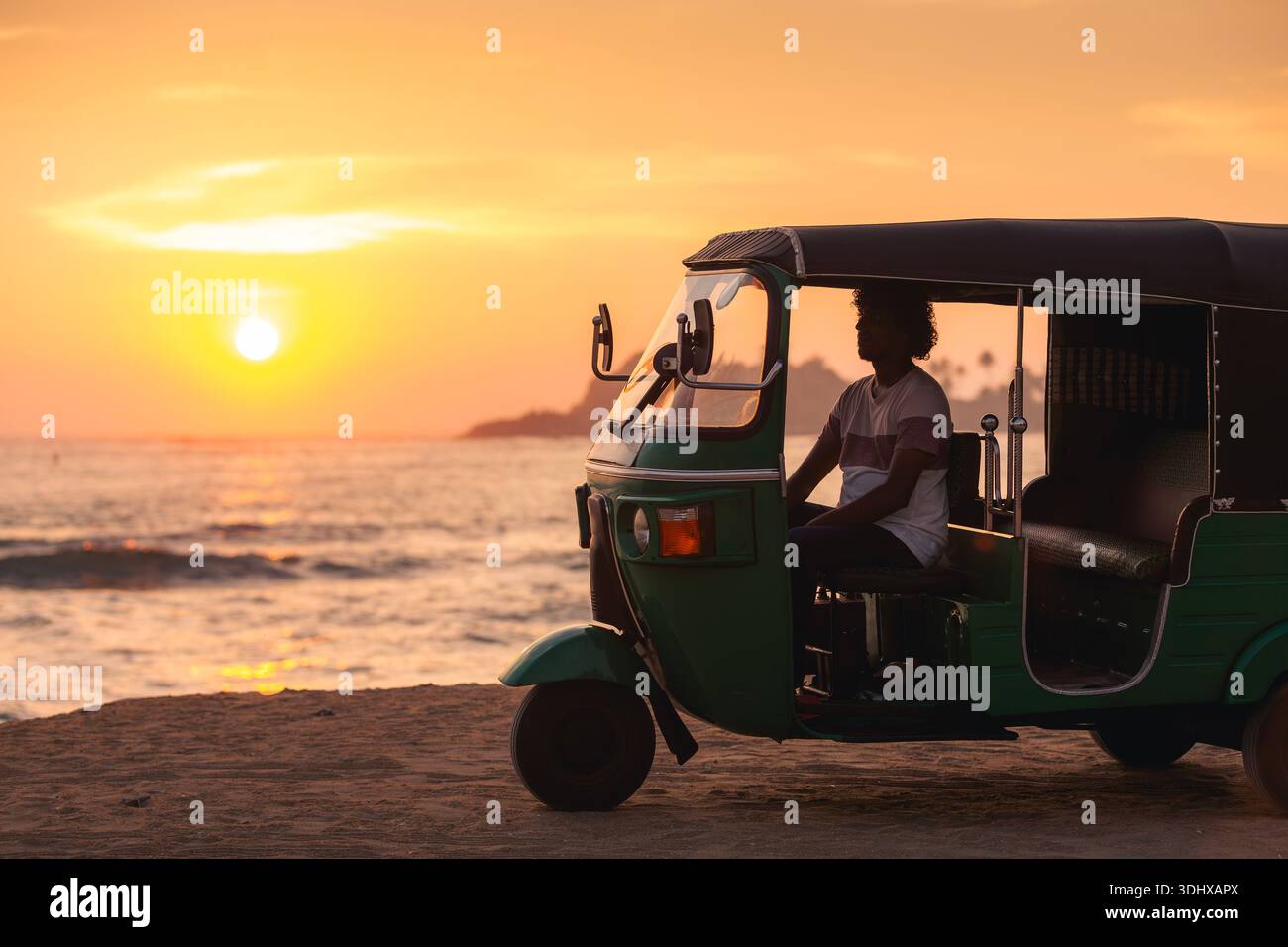Chauffeur de tuk tuk local attendant à l'intérieur du véhicule sur la plage de sable fin pendant le coucher du soleil sur l'océan au Sri Lanka. Lumière chaude du soir et atmosphère tropicale calme. Banque D'Images