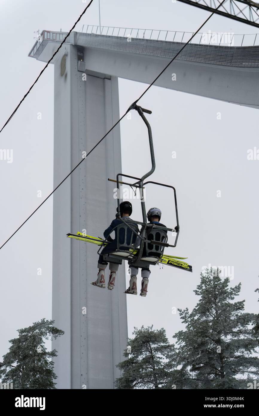 Lahti, Finlande - 18 janvier 2025 - deux sauteurs à ski montant un télésiège avec la grande colline de Lahti en arrière-plan par une journée brumeuse Banque D'Images