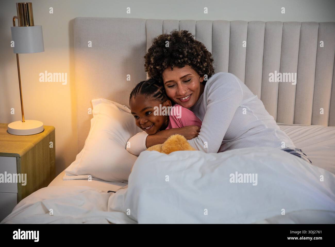 Mère et fille allongées ensemble dans la chambre sur le lit serrant sous couette blanche près de la lampe de chevet. Famille, intimité, chaleur, confort, coucher, affection, soins || autorisation du modèle Banque D'Images