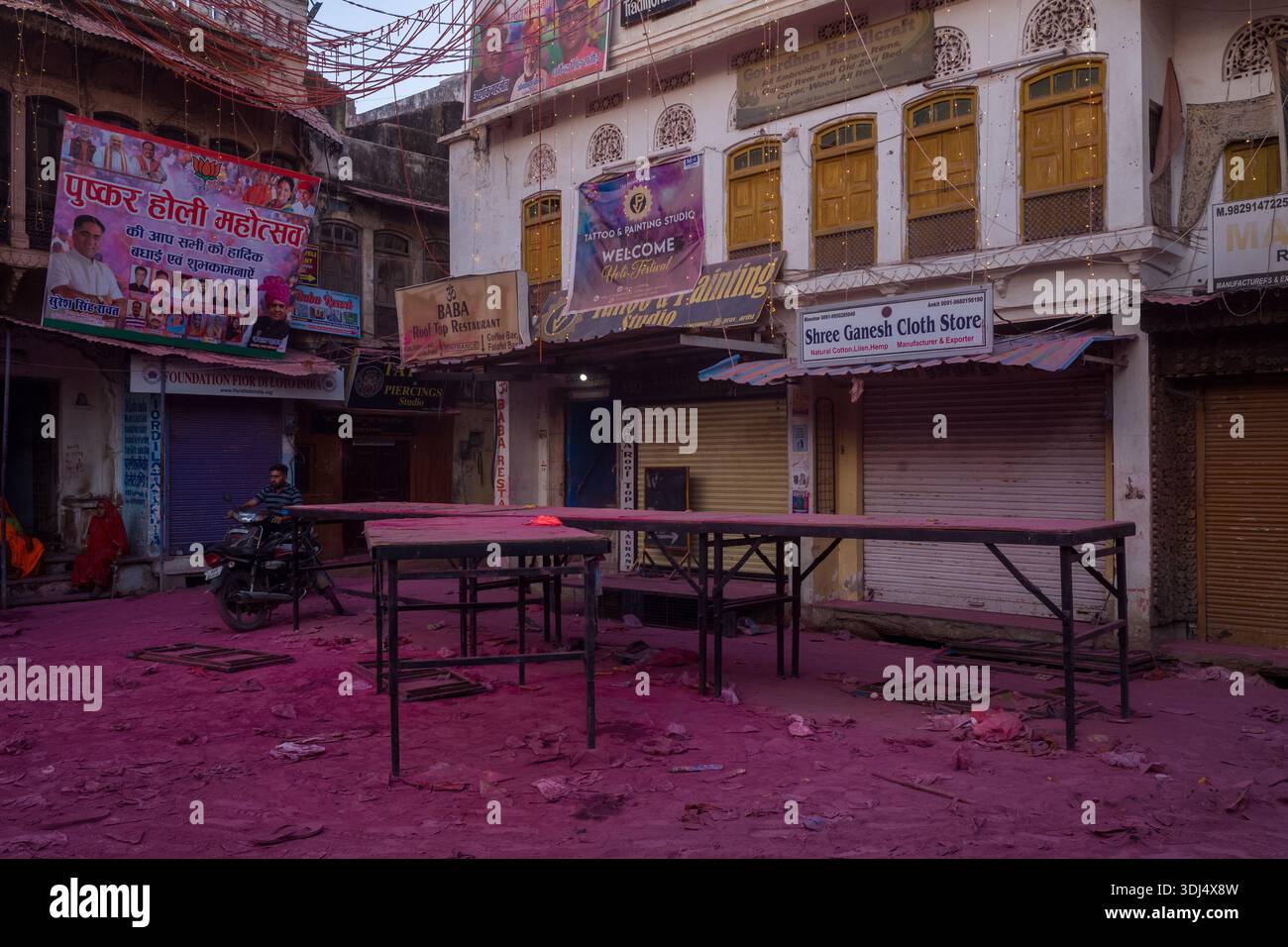 Pushkar, Inde - 18 mars 2022 : plusieurs magasins fermés avec des enseignes visibles et des bannières sont vus le long d'une rue couverte de poudre rose Holi, avec des tables vides installées au premier plan et un homme assis sur une moto. Banque D'Images