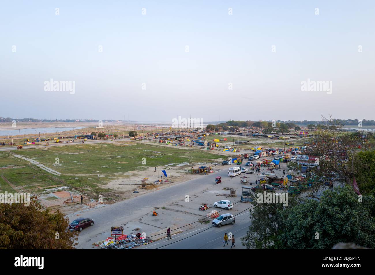 Prayagraj, Inde - 21 mars 2024 : L'image montre une vue aérienne d'un grand terrain de parade ouvert avec des taches d'herbe, des véhicules dispersés, des groupes de personnes, des stalles temporaires, des vendeurs de bord de route, et la rive de la rivière en arrière-plan sous un ciel clair pendant la journée. Banque D'Images
