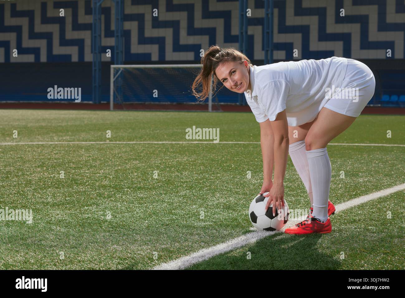 Joueuse de football confiante en uniforme blanc se prépare pour un match sur un terrain extérieur, souriant à la caméra. Banque D'Images