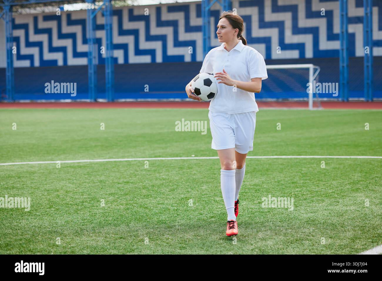 Joueuse de football féminine confiante en uniforme blanc marchant à travers le champ avec la balle, prête pour l'entraînement ou le jeu. Banque D'Images