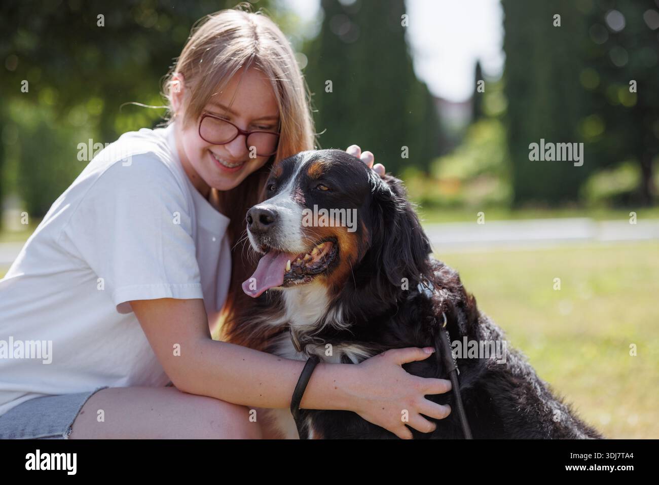 Une fille blonde caresse son chien de montagne bernois dans un parc ensoleillé, montrant leur lien affectueux et le plaisir du moment Banque D'Images