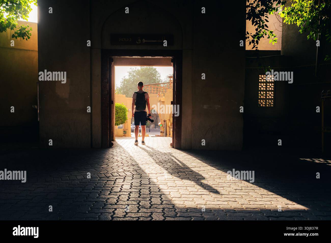Touriste tenant caméra marchant à travers la porte du souk traditionnel dans la ville historique de Nizwa en Oman. Forte lumière matinale projetant une longue ombre, voyage ex Banque D'Images
