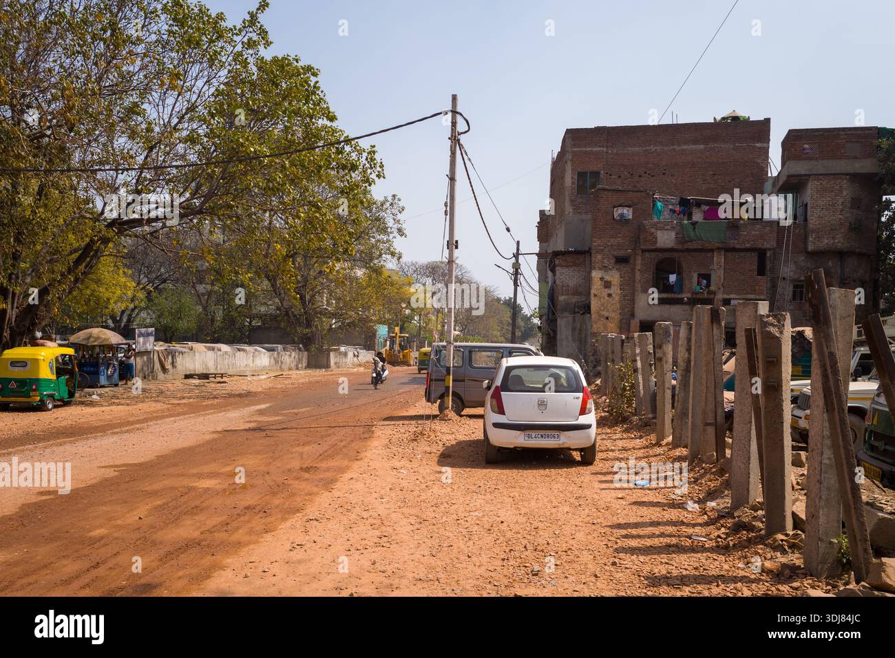 Delhi, Inde - 13 mars 2022 : une rue non pavée dans la région de Karol Bagh montre des voitures garées, un pousse-pousse automobile, un motocycliste, des poteaux électriques avec des fils aériens et des bâtiments résidentiels en briques à plusieurs étages avec du linge suspendu aux balcons pendant la journée. Banque D'Images