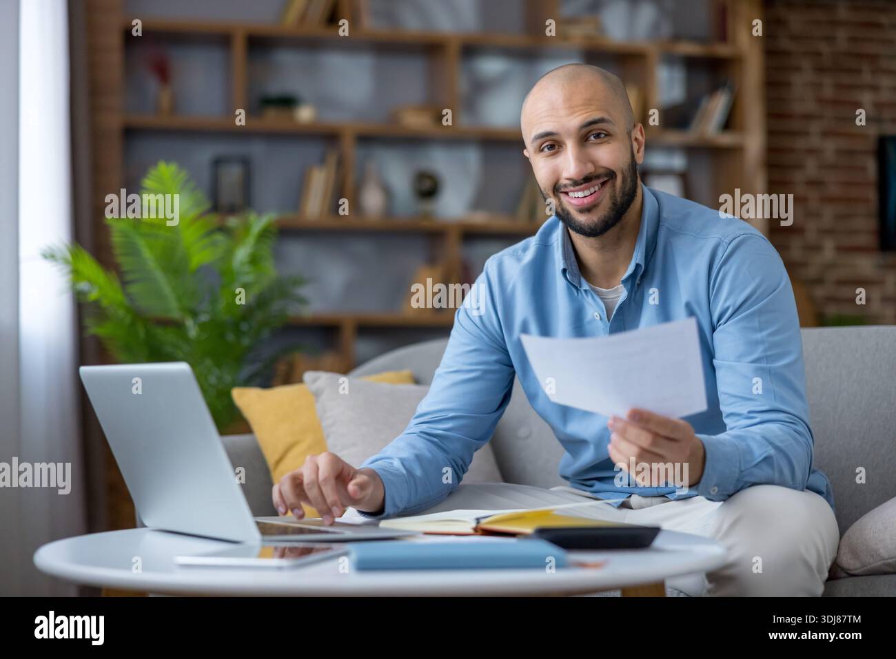 Jeune homme souriant à la caméra tout en travaillant sur un ordinateur portable, tenant des documents, calculant le budget et gérant les finances à partir de la configuration du bureau à domicile dans un salon confortable Banque D'Images
