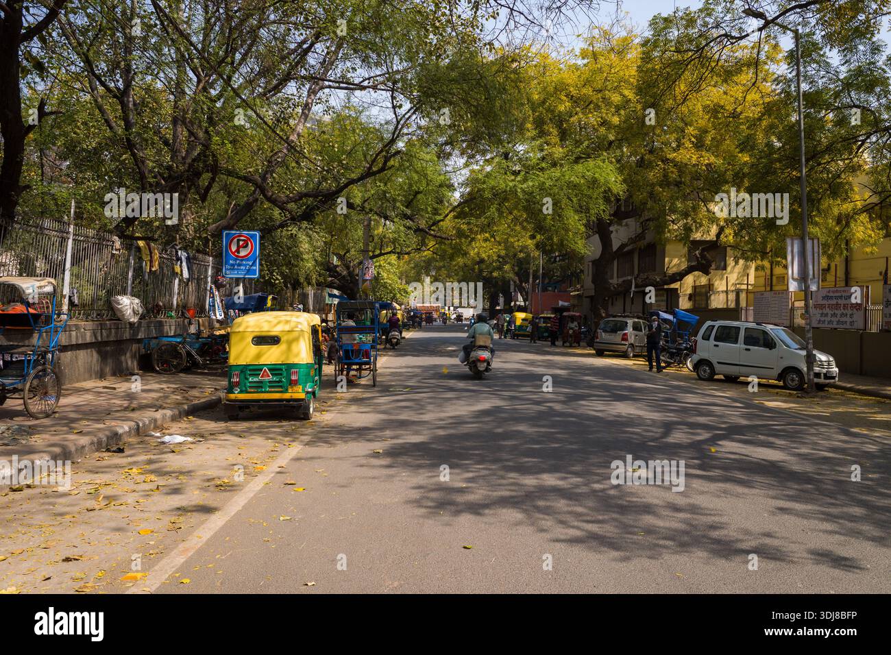 Delhi, Inde - 13 mars 2022 : une rue dans le quartier de Karol Bagh montre des pousse-pousse automobiles, des pousse-pousse à vélo, des véhicules stationnés, des piétons et de grands arbres feuillus projetant des ombres sur la route pendant la journée. Banque D'Images