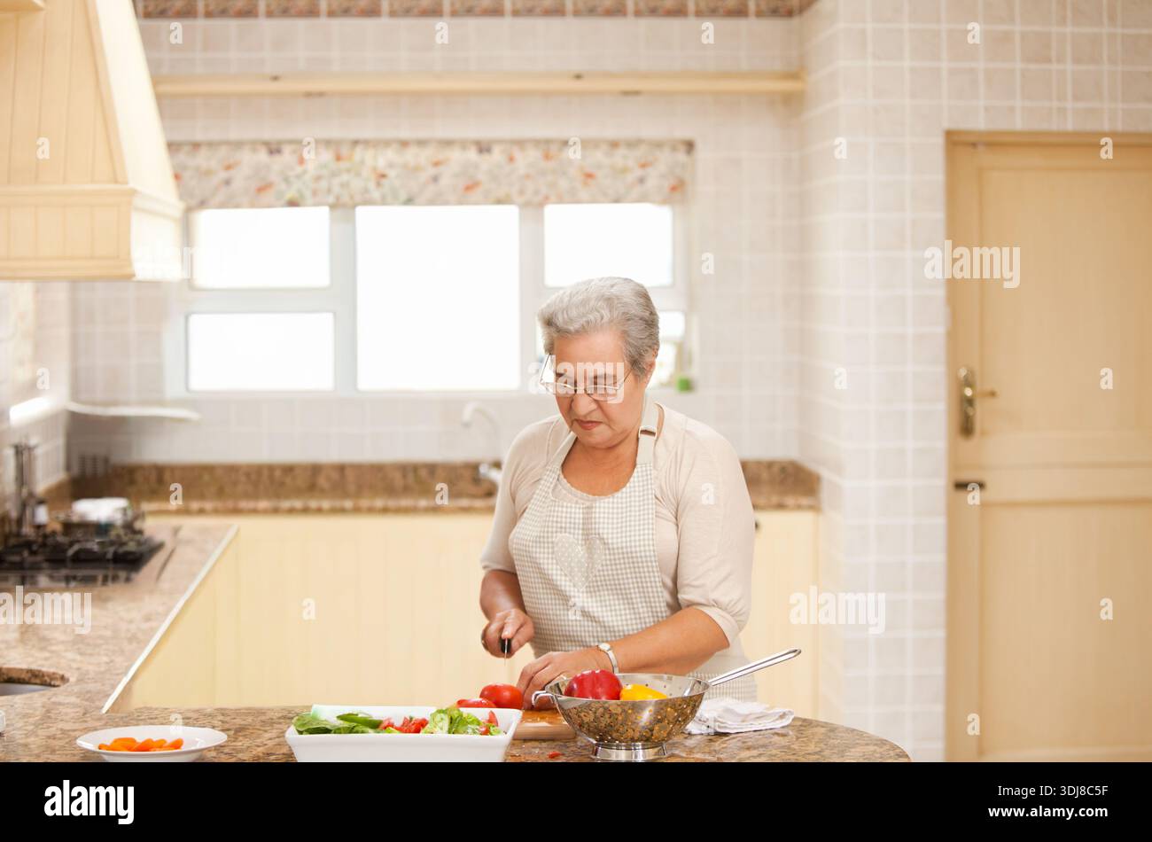 Femme senior hachant des tomates et mélangeant la salade sur l'île de granit portant un tablier à carreaux avec un couteau. Cuisine, ensoleillé, confortable, homecooking, légumes, sl Banque D'Images