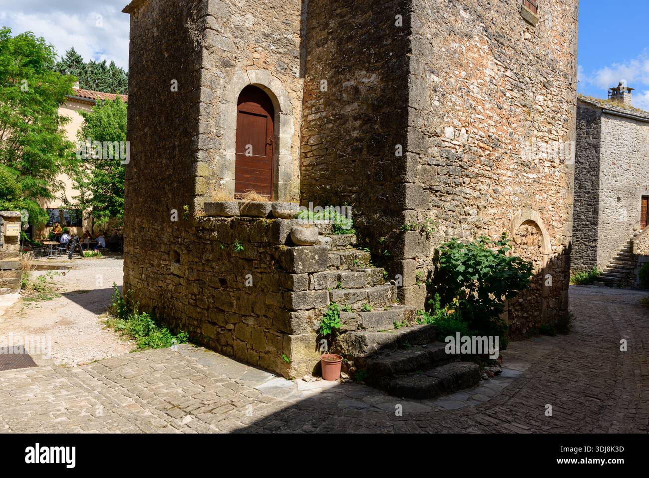 La Couvertoirade, France - 26 juin 2022 : un bâtiment historique en pierre avec une entrée surélevée et un escalier extérieur en pierre se dresse à une intersection de pavés dans le village médiéval, avec de la verdure et des gens assis à des tables extérieures visibles en arrière-plan. Banque D'Images
