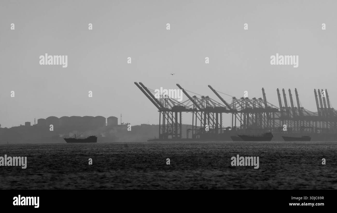 Une photo frappante en noir et blanc d'un port industriel de Salalah, Oman, montrant les silhouettes de grandes grues à conteneurs et de cargos contre un h. Banque D'Images