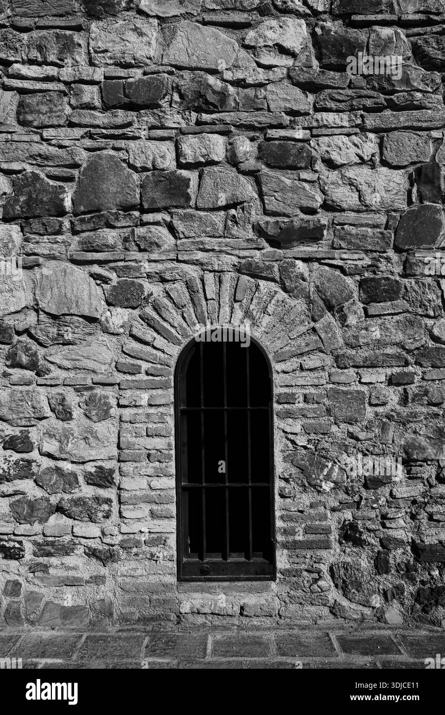 Une photographie en noir et blanc très contrastée d'un ancien mur de pierre avec une fenêtre centrale voûtée avec des barres de fer foncé. La texture rugueuse Banque D'Images