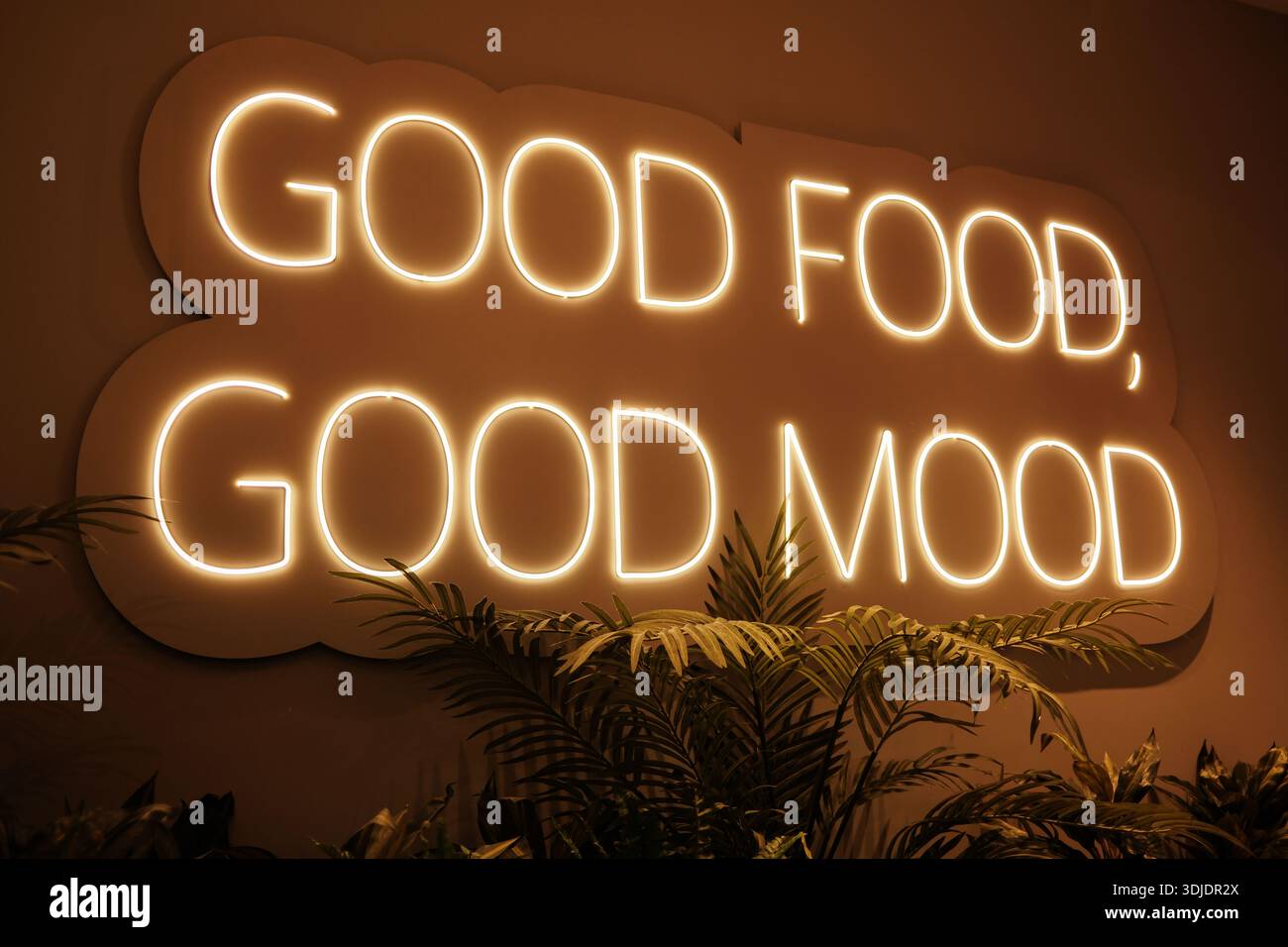 Panneau mural au néon chaud lisant Good Food Good Mood briller dans un intérieur confortable avec des plantes, créant une atmosphère détendue liée à la salle à manger, le confort et Banque D'Images