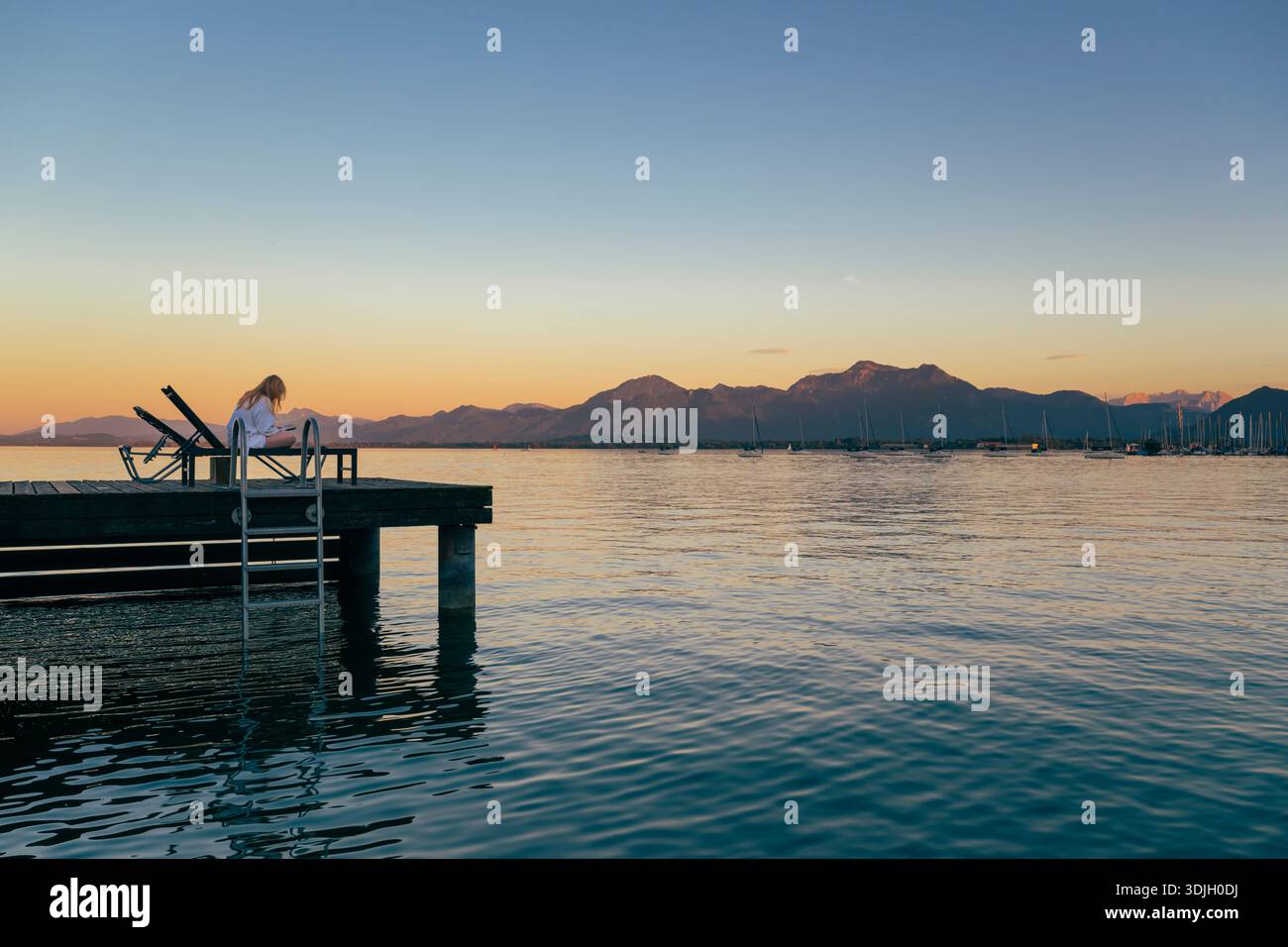 Une jeune femme en peignoir blanc se trouve sur un transat sur une jetée au lac Chiemsee dans le crépuscule incandescent, lisant un livre, Bavière, Allemagne Banque D'Images