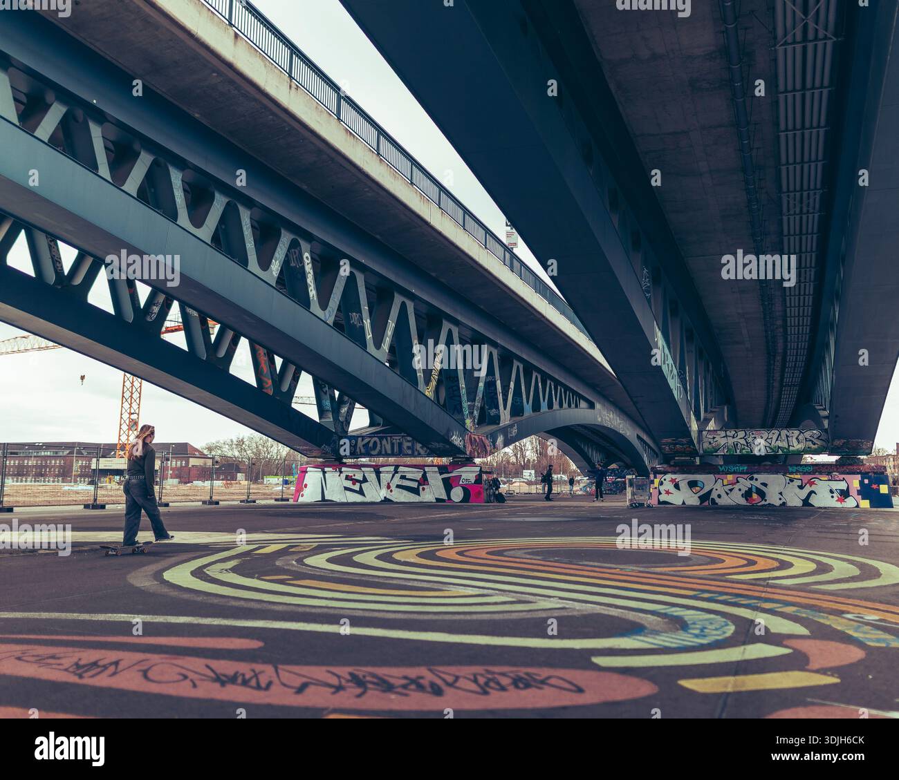 Scène urbaine de skateboard sous un pont arqué moderne, avec des arts circulaires colorés au sol, des skateurs en mouvement et une ville industrielle en toile de fond sur un Banque D'Images