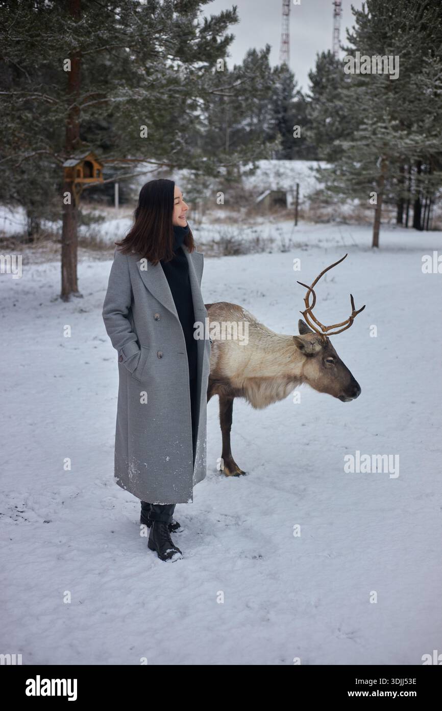 femme dans un long manteau se tient à côté d'un renne dans une zone enneigée entourée d'arbres. scène montre le temps froid et la présence de la faune dans le w Banque D'Images