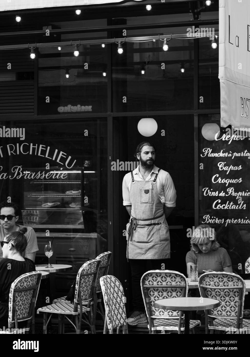 Un homme dans un tablier debout à l'extérieur de son café pendant que les clients s'assoient et mangent. Banque D'Images