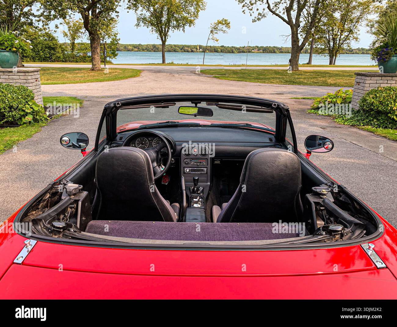 Le cabriolet rouge vif avec la capote abaissée révèle des sièges en cuir noir et un tableau de bord poli. Conduire en plein air près d'un lac crée une vaca insouciante Banque D'Images