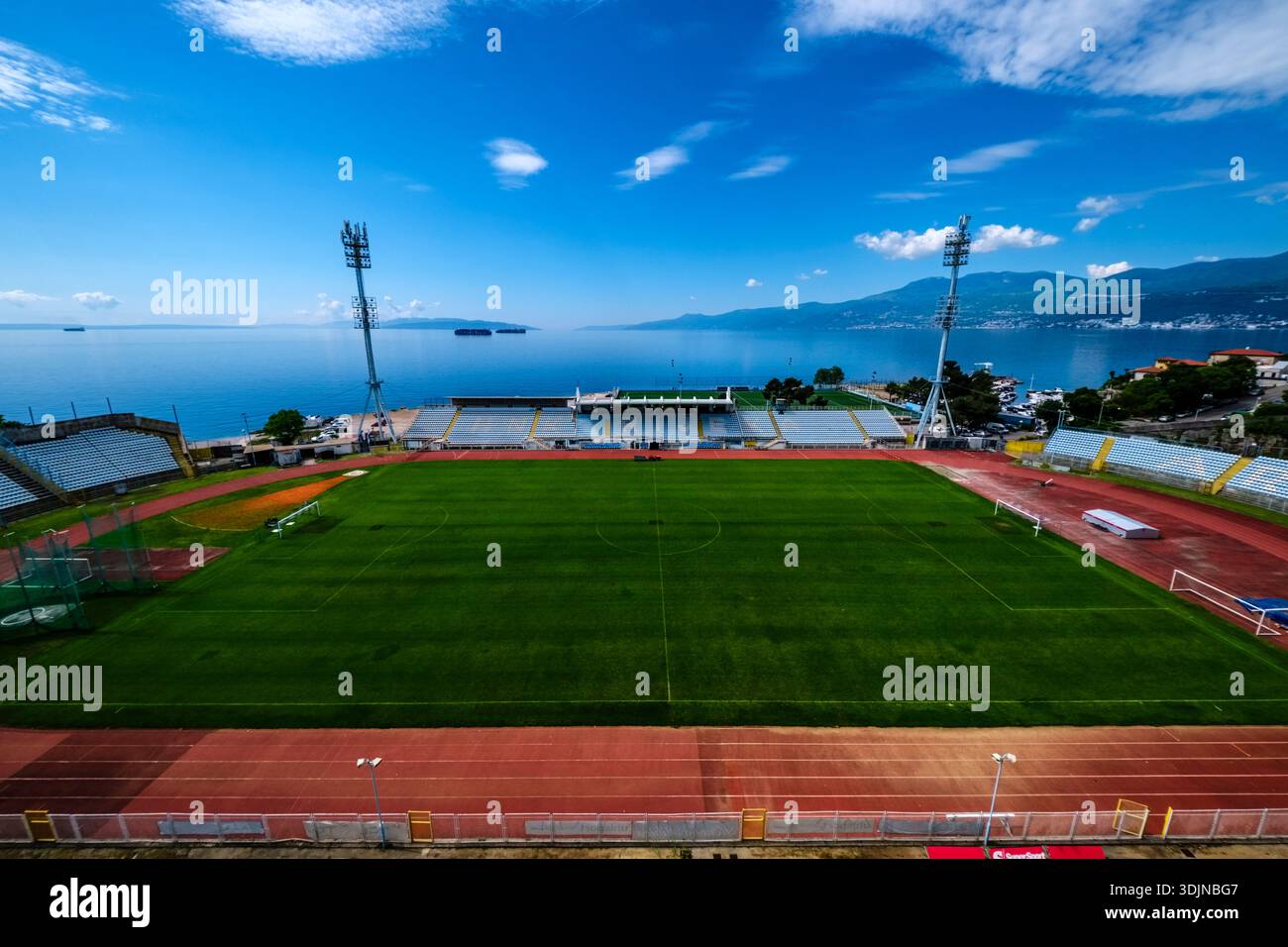 Stade Kantrida avec terrain vert et projecteurs au bord de la mer Adriatique. Učka chaîne de montagnes au loin sous un ciel bleu. Banque D'Images
