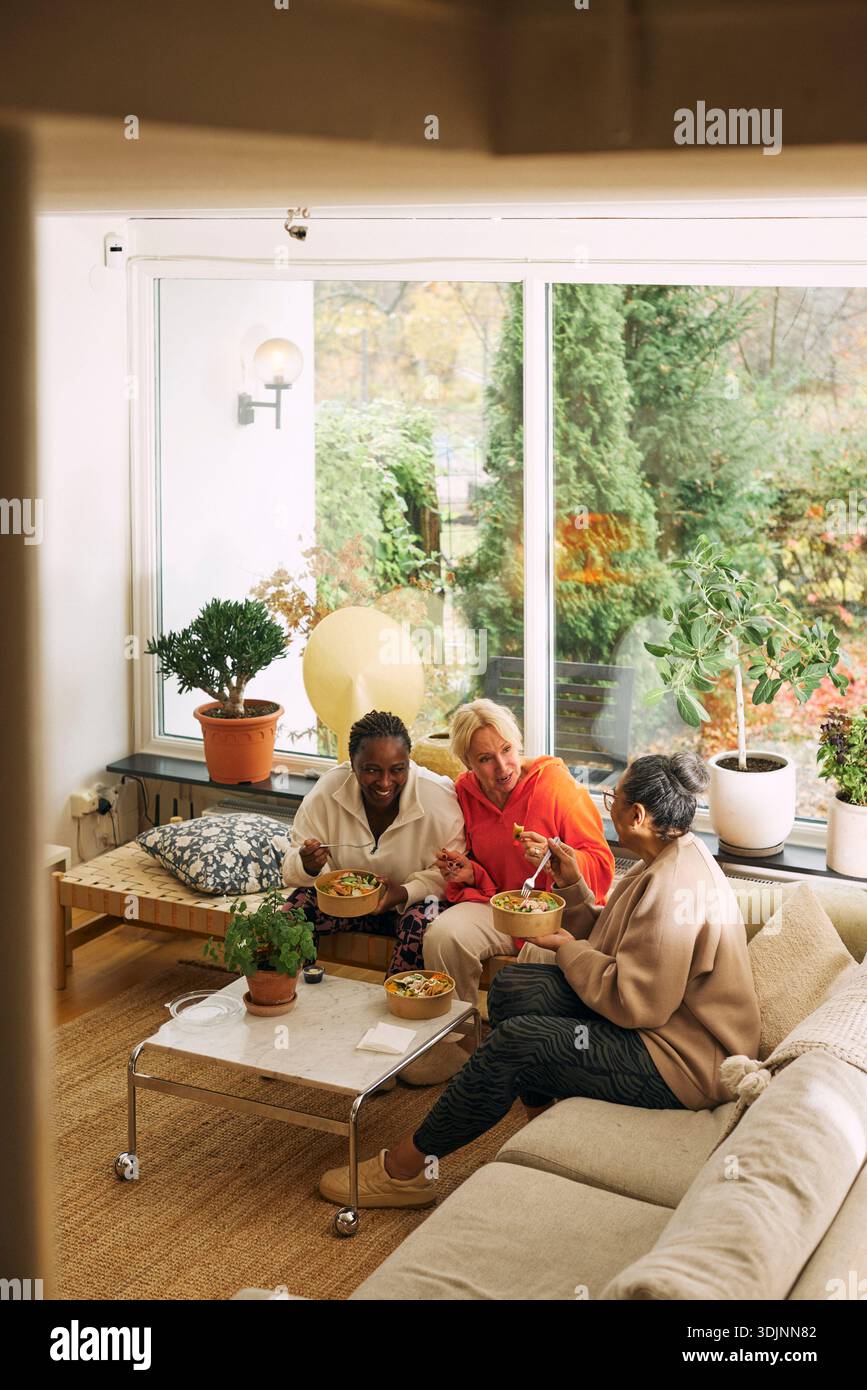 Vue en angle élevé d'amies féminines multiraciales mangeant le déjeuner tout en étant assis sur le canapé à la maison Banque D'Images