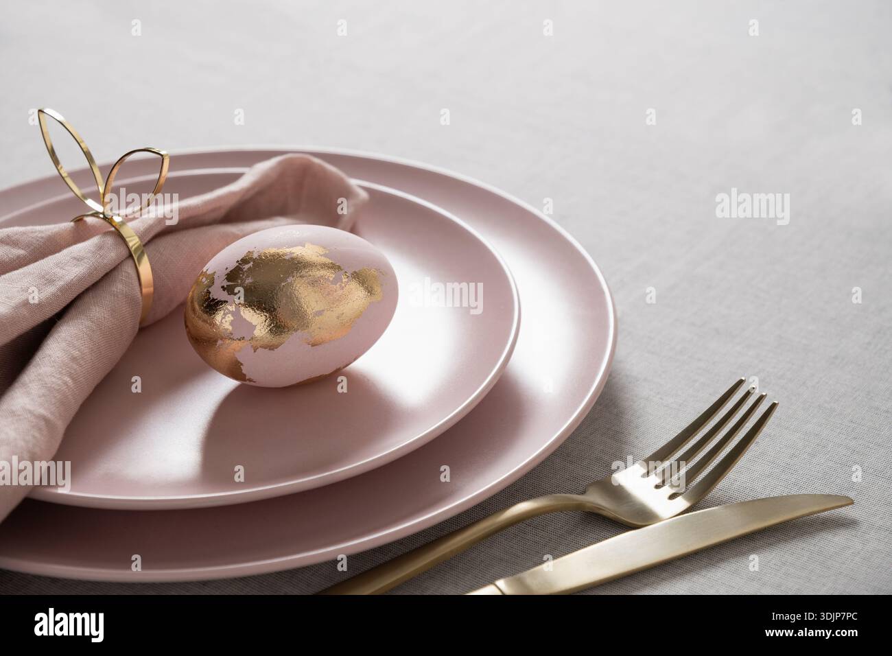 Élégante table de Pâques avec assiettes rose pastel, oeuf décoratif avec feuille d'or et couverts dorés sur nappe en lin léger. Vacances de printemps luxueuses Banque D'Images