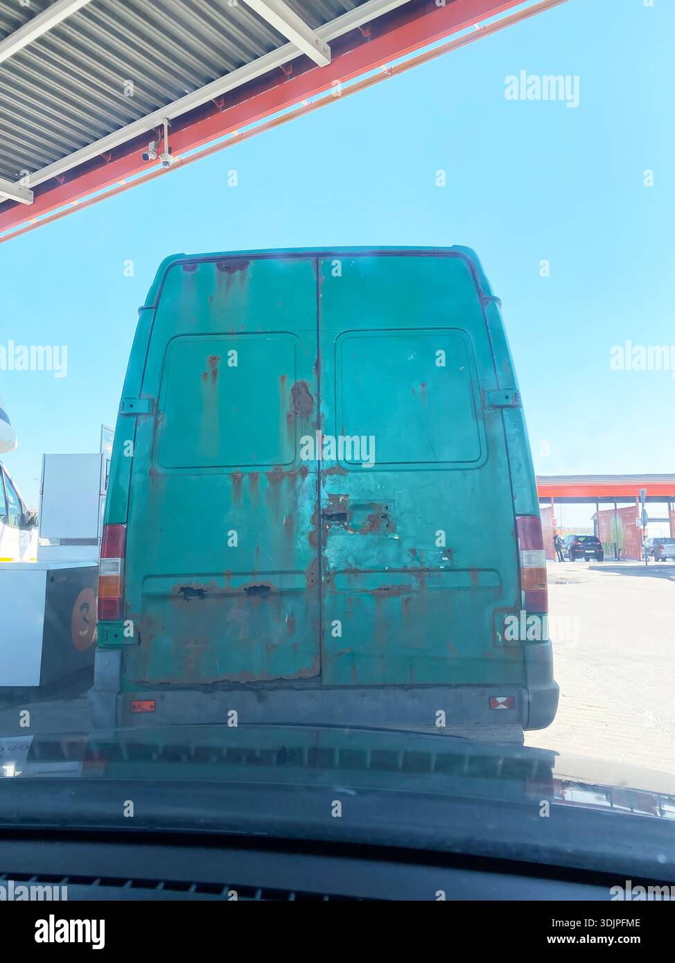 Le fourgon de livraison vert avec des taches de rouille est garé à une station-service, des pompes à carburant et un ciel bleu clair visible en arrière-plan pendant les heures de jour Banque D'Images