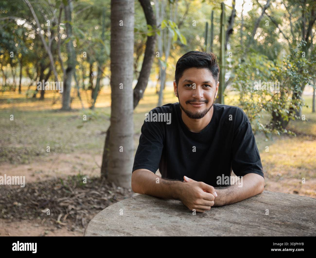 Image d'un jeune homme appuyé sur une table de pierre dans un parc, regardant la caméra avec une expression amicale. Photographie adaptée aux concepts de bien-être Banque D'Images