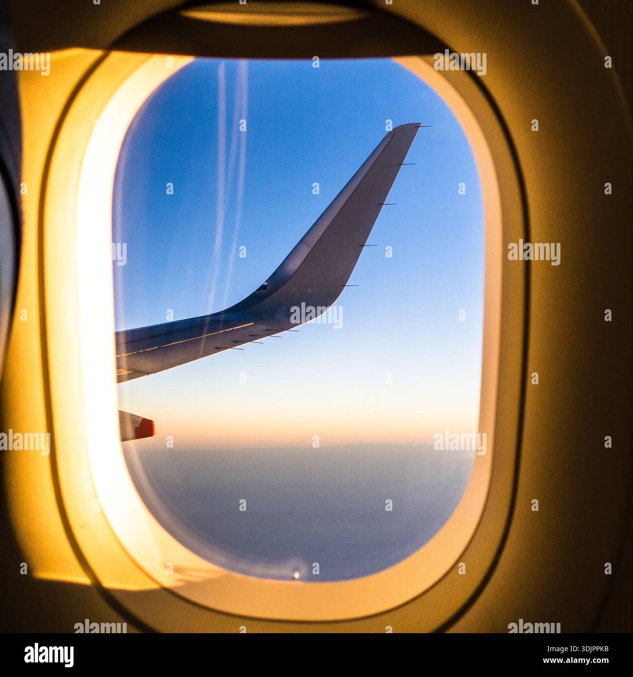Aile d'avion vue de la fenêtre du passager pendant le vol au coucher du soleil, avec un éclairage de bord doré contre le ciel bleu et les nuages lointains Banque D'Images