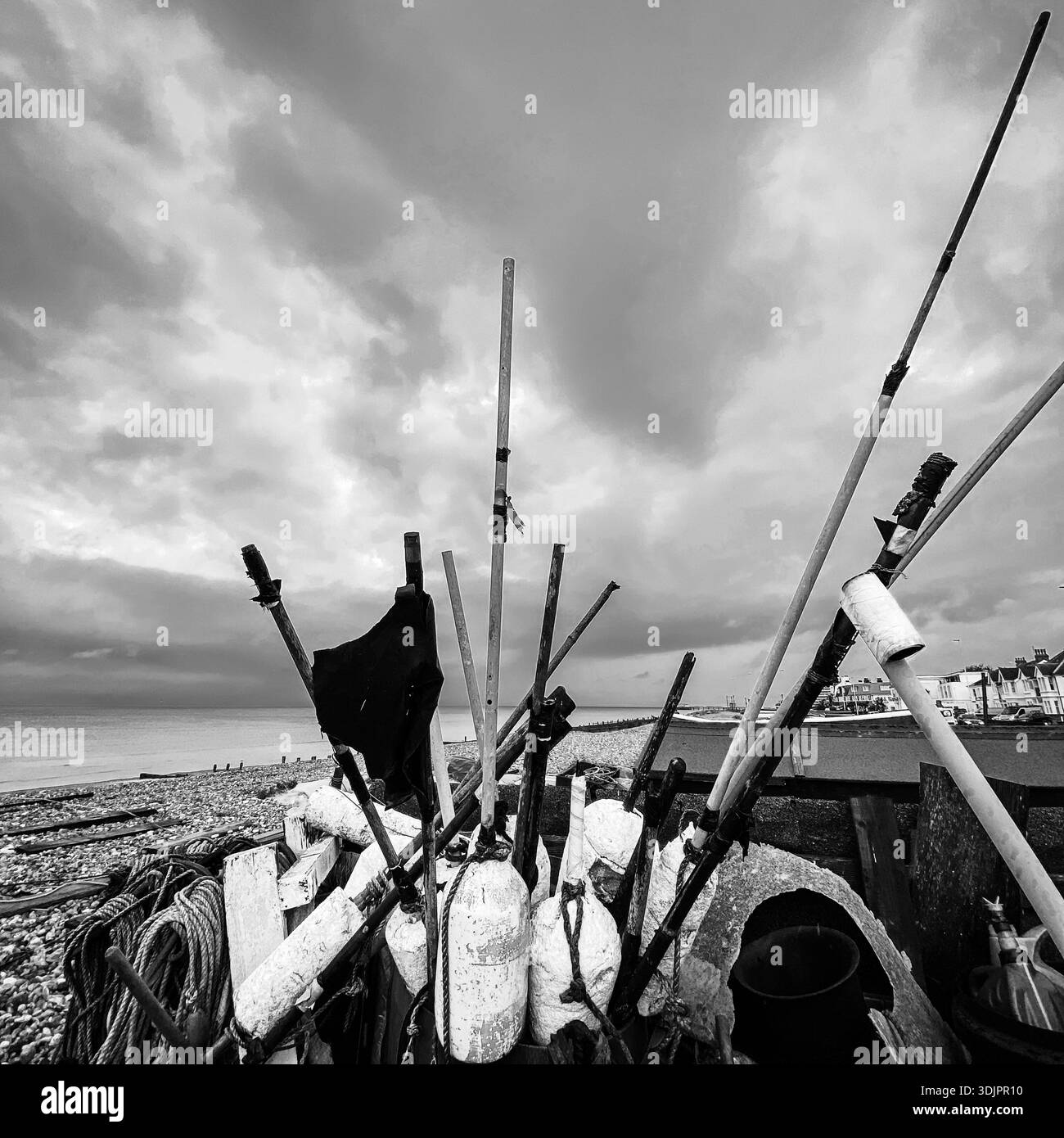 Gros plan de cannes à pêche droites, filets, bouées et engins sur un bateau de plage de galets en monochrome, avec un ciel orageux et un quai au bord de la mer anglaise. - Image de stock capturée avec un smartphone